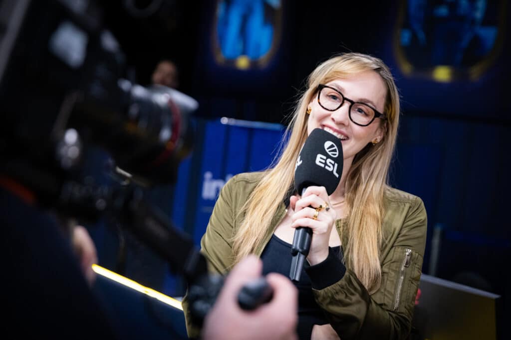 Sjokz on camera at ESL IEM Katowice 2025. She is pictured holding an ESL mic while reporting on stage.