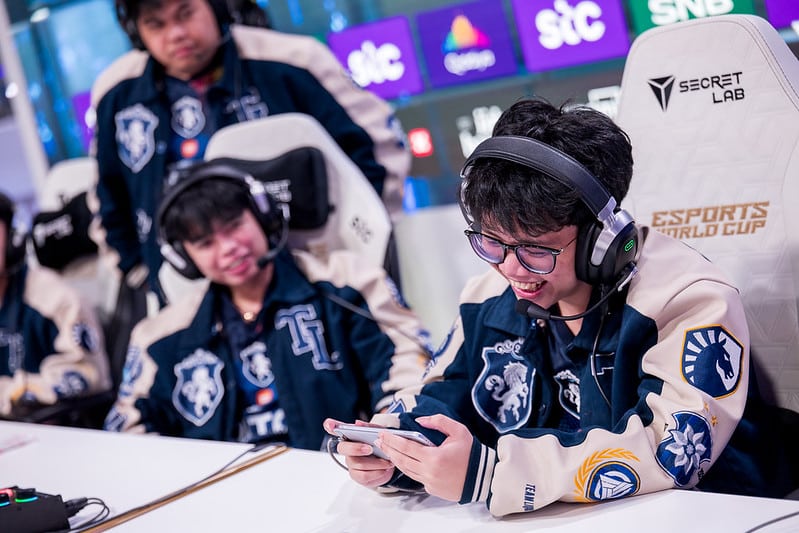 MLBB at the Esports World Cup - a player holds their competition phone while smiling. Two of their teammates watch in the background