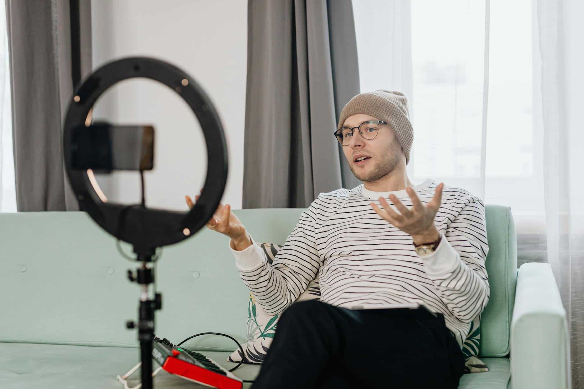 Man with glasses and beanie livestreaming on a couch with a ring light and smartphone.