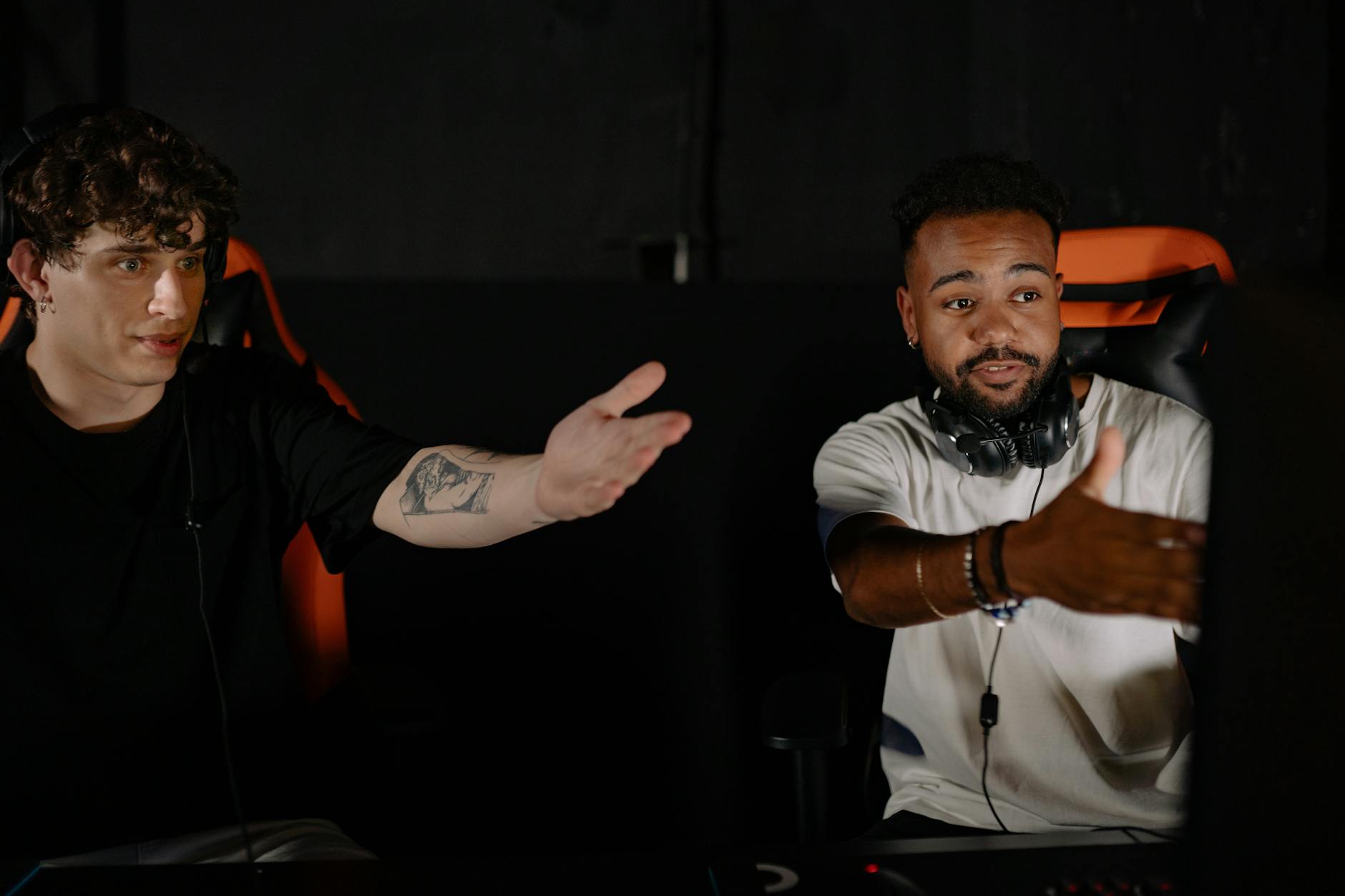 Two men sitting in gaming chairs discussing while pointing at computer screens.