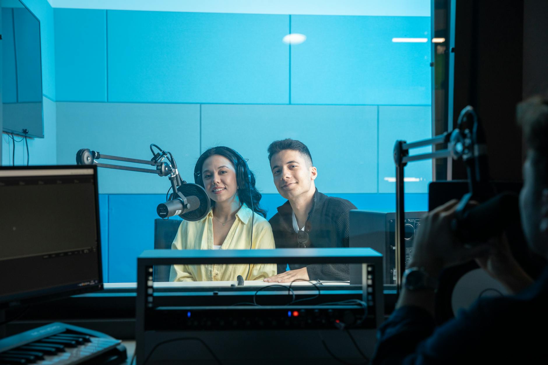 Two people in a broadcast studio with microphones and headphones, seen through a window.