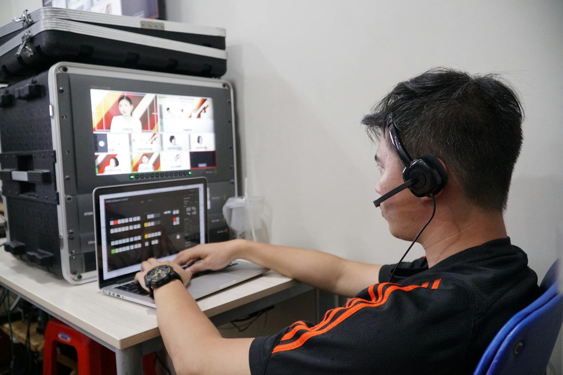 Man with headset using laptop for live streaming, with broadcast screens in the background.