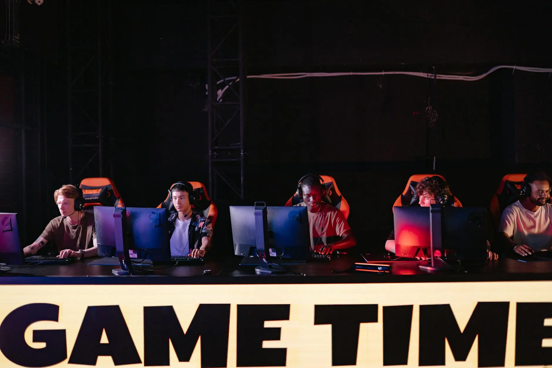 Five gamers seated behind computers in a dark esports arena with vibrant lighting.
