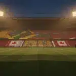 Floodlit stadium at golden-hour with packed stands in yellow-green and red-white colours, empty pitch, dramatic shadows and lighting.
