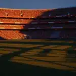 Floodlit football stadium with packed crowd at twilight, empty pitch with dramatic shadows, warm golden and cool blue lighting.
