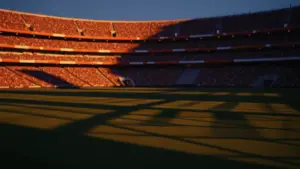 Floodlit football stadium with packed crowd at twilight, empty pitch with dramatic shadows, warm golden and cool blue lighting.
