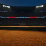 Floodlit stadium at golden hour with packed stands and dramatic shadow patterns on empty pitch.